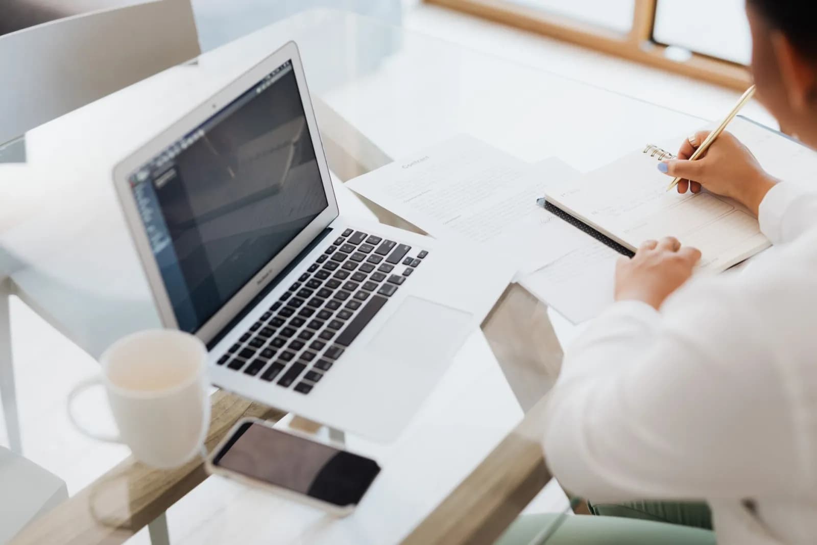 A laptop, notebook, and coffee mug on a glass desk